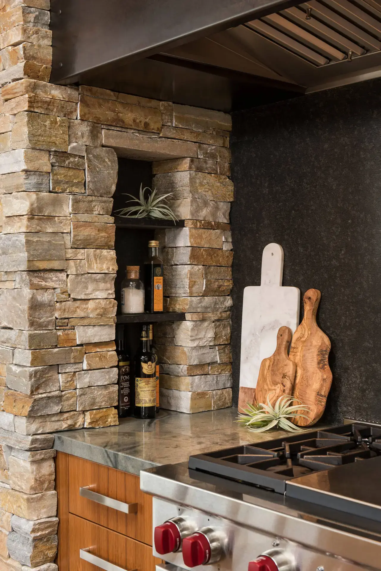 Modern kitchen featuring luxury custom cabinetry with a stone backsplash, integrated shelving for bottles and spices, and wooden and marble cutting boards displayed on a sleek countertop adjacent to a stainless steel stove.