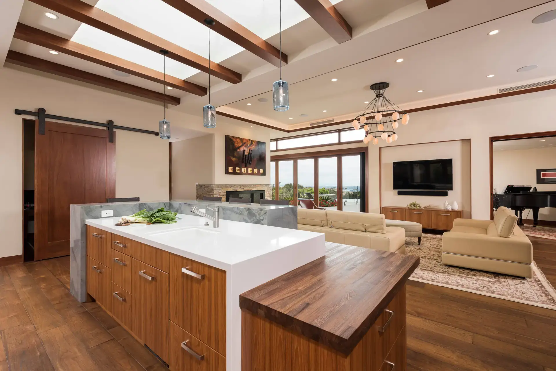 Luxurious open-concept living room and kitchen with custom wood cabinetry, modern lighting fixtures, beige sofas, and large windows offering a scenic view. High-end interior design in San Diego and LA.