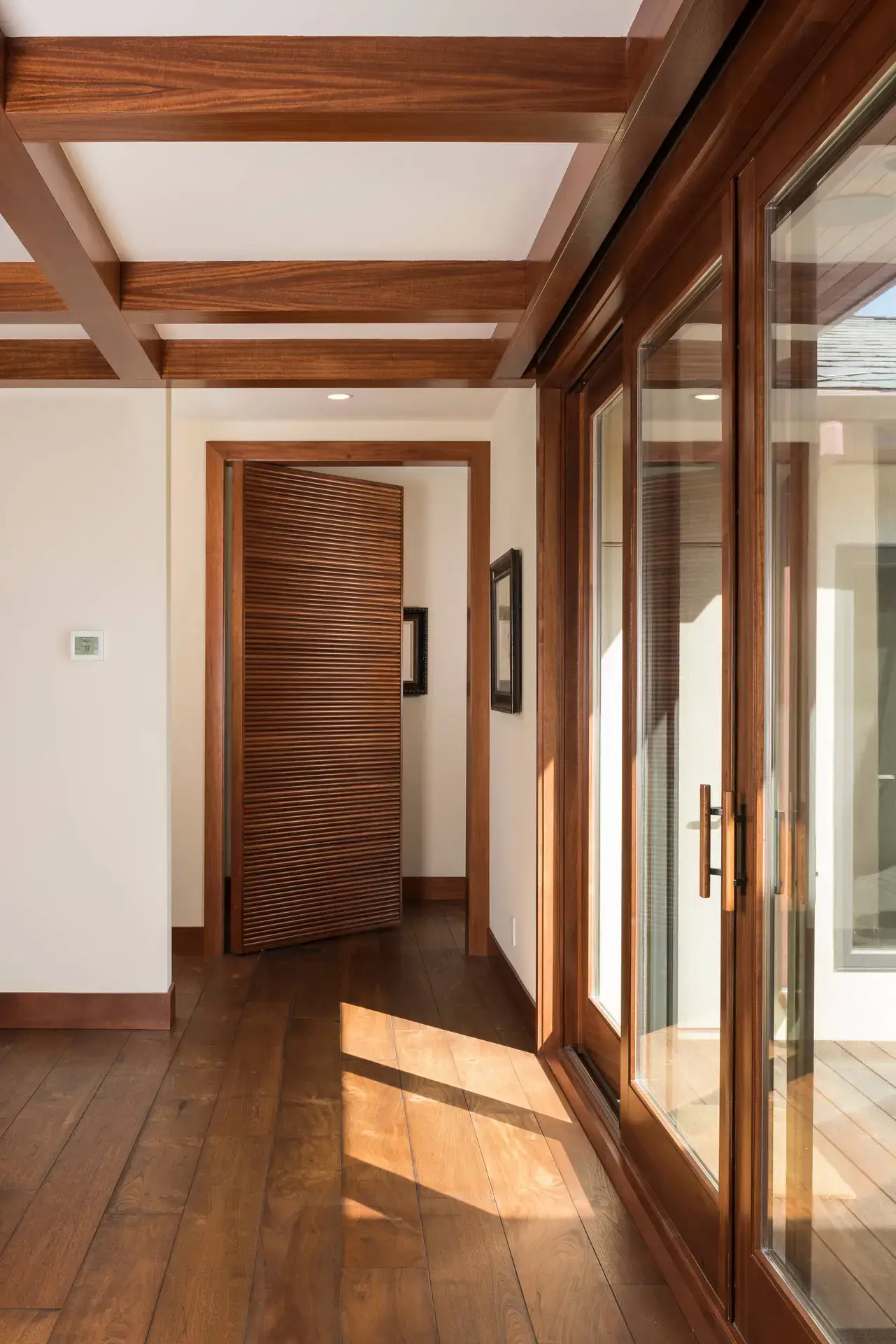 Wide-angle view of a modern interior hallway featuring polished wooden floors, ceiling beams, and a partially open louvered wooden door. Sunlight streams throu