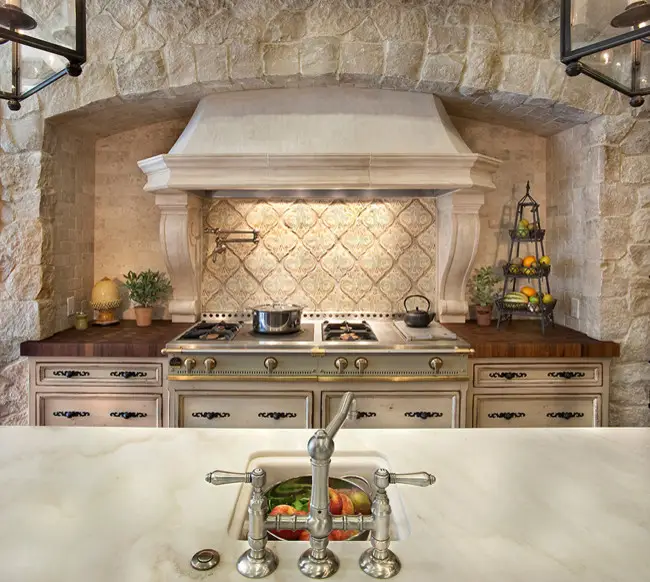 Elegant kitchen with custom cabinetry, featuring a white stove hood, ornate tile backsplash, stone walls, and a wooden countertop, enhancing modern kitchen remodel and storage design.