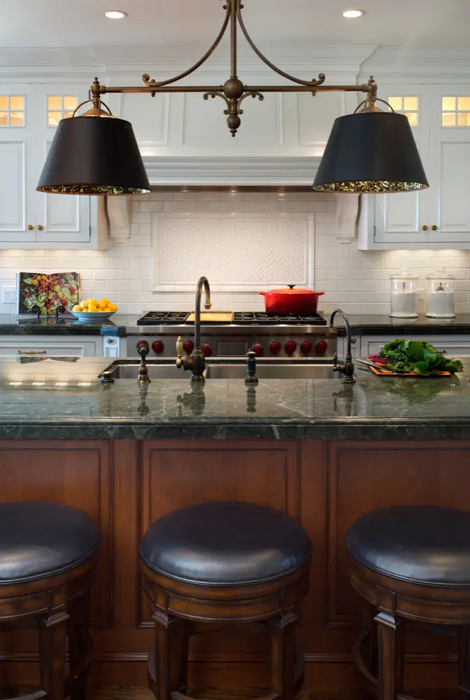 Modern kitchen with elegant custom cabinets, featuring a green marble countertop island, bronze fixtures, and black pendant lights. White cabinetry and a classic stovetop with a red pot add sophistication to the design.