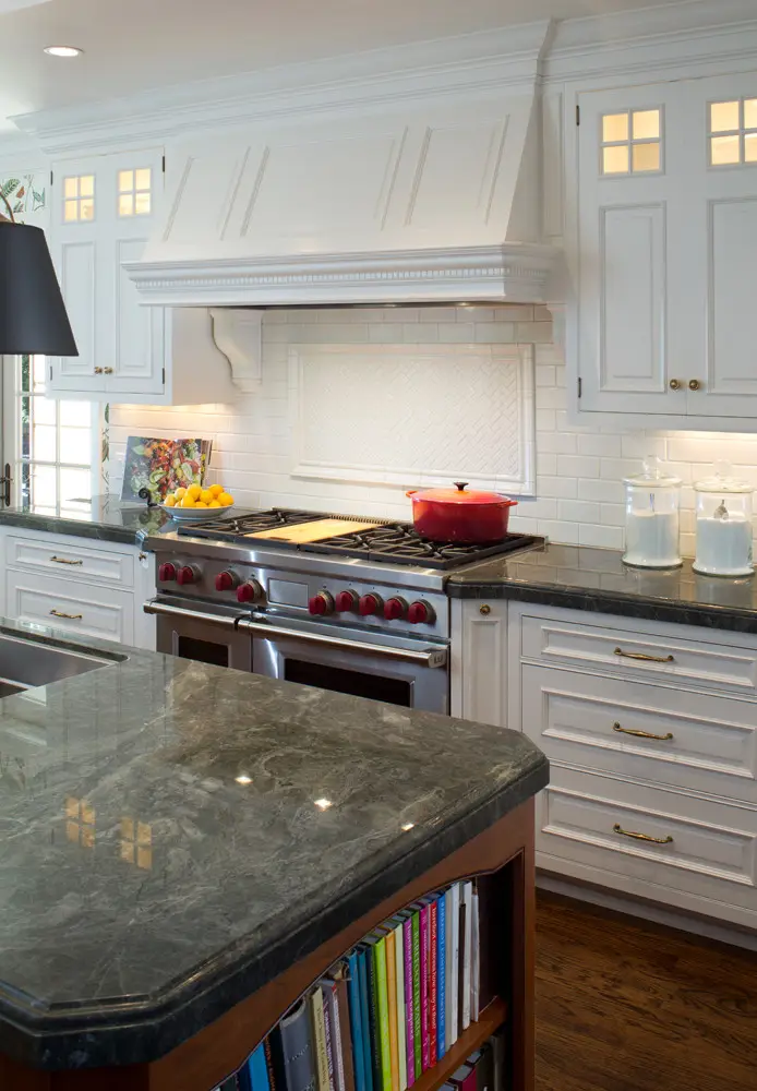 Modern kitchen with elegant custom white cabinets, granite countertops, and a stainless steel oven with red knobs. Features a sophisticated backsplash, a center island with dark countertop, and an integrated bookcase.