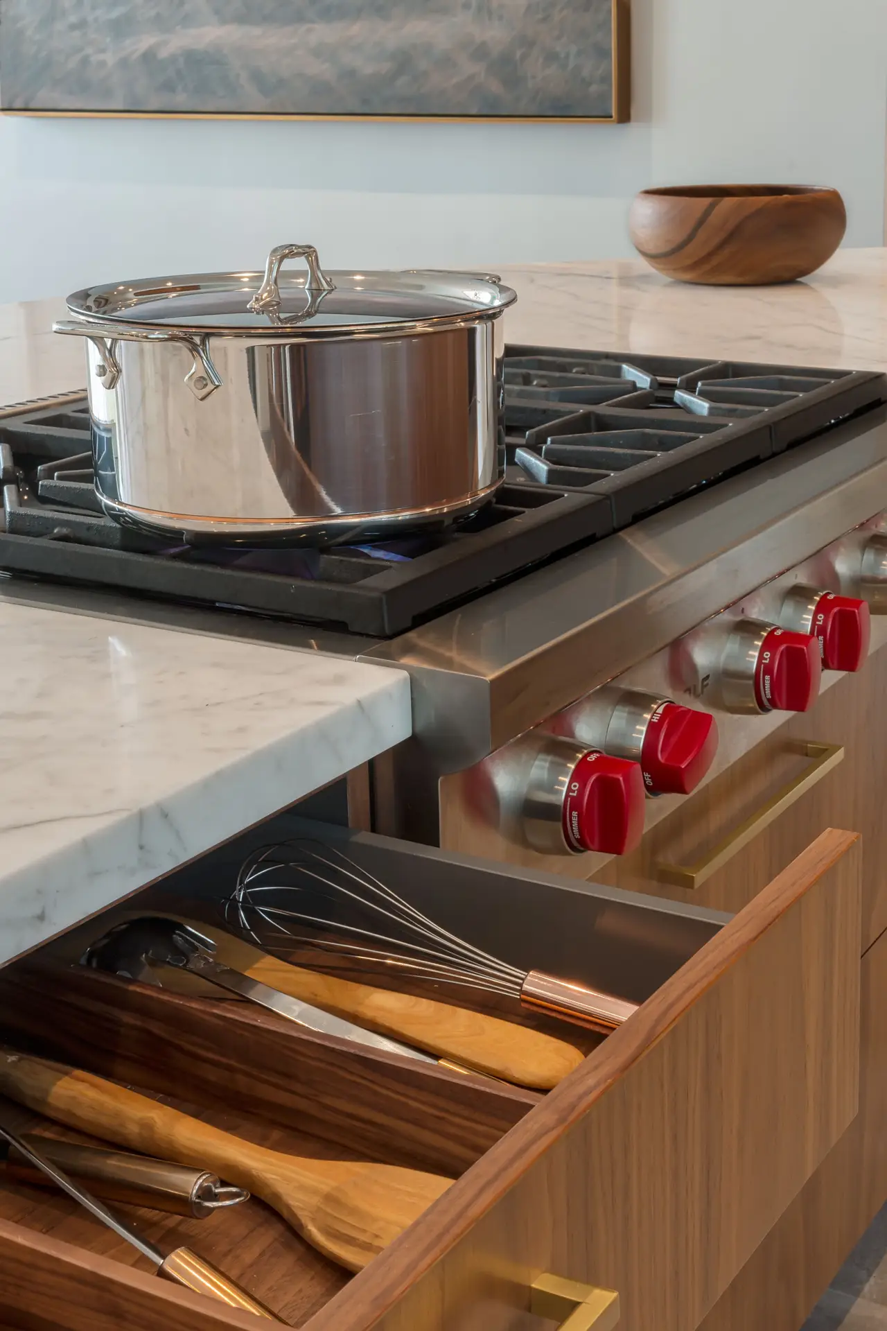 Luxurious kitchen with custom cabinetry, featuring a stainless steel pot on a modern stovetop, red knobs, and an elegant marble countertop. A wooden utensil drawer is open, showcasing high-end kitchen tools, emphasizing stylish storage solutions.