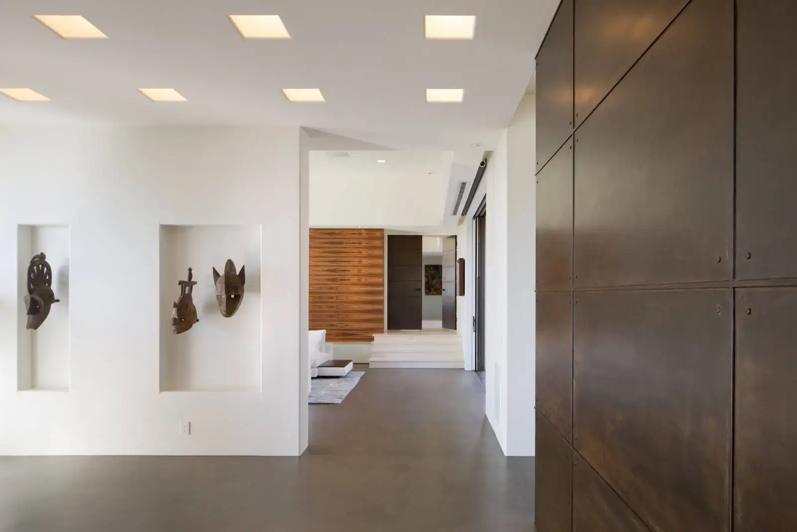 Modern interior with minimalist design featuring recessed lighting and wall niches displaying wooden sculptures. The space includes smooth concrete flooring and dark wooden panels, reflecting luxury European kitchen and interior solutions.