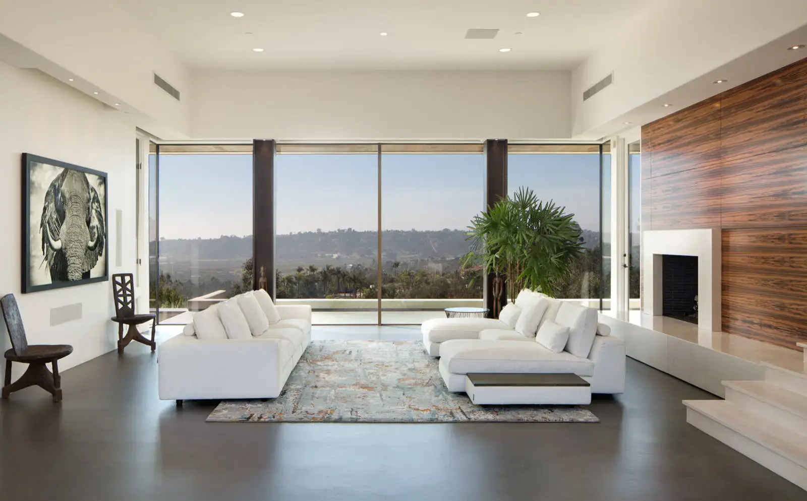 Spacious modern living room featuring white sofas, a large plant, artistic wall decor, and a wide glass window offering panoramic views. The room includes a sleek wooden accent wall with a fireplace, emphasizing luxury and contemporary interior design.