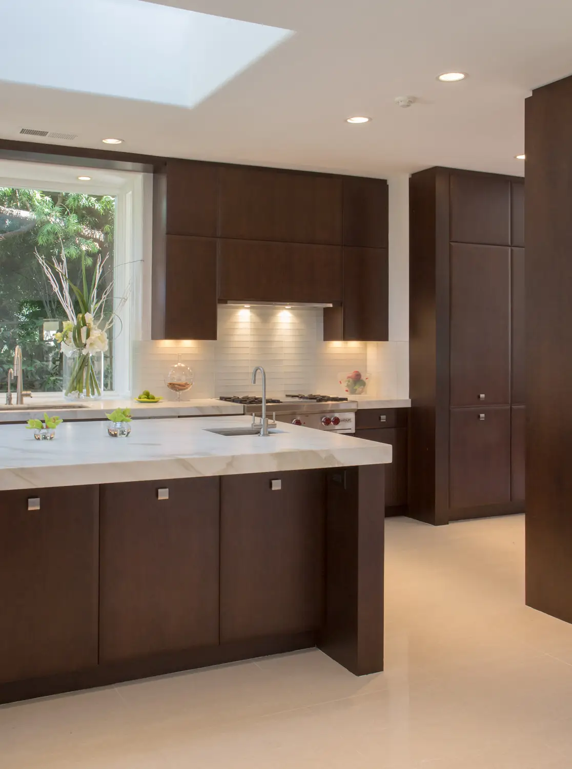 Modern luxury kitchen featuring custom dark wood cabinetry, a sleek marble countertop, and a built-in stove with under-cabinet lighting. Large window with garden view and decorative vases with flowers.