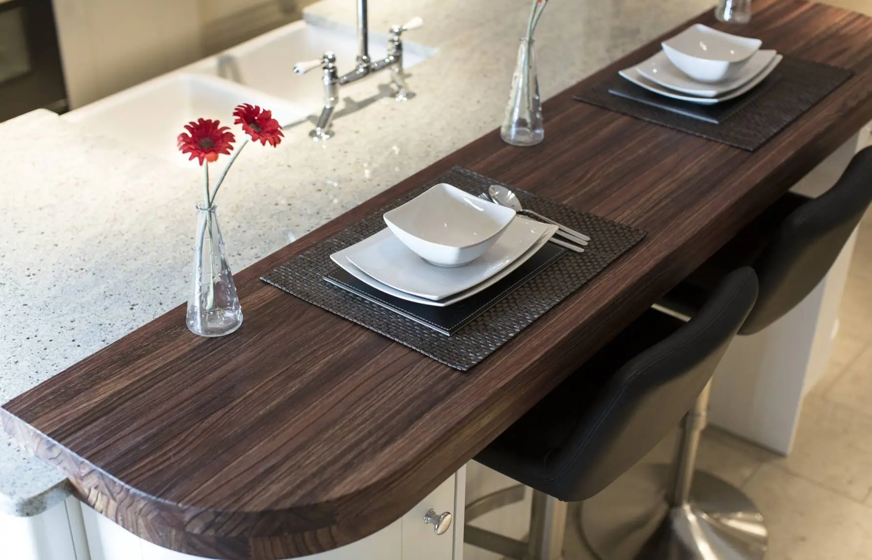 Elegantly designed kitchen island with dark wood surface, featuring white dinnerware, black placemats, and modern barstools, complemented by minimalist vases with red flowers. Ideal for luxury home decor and custom cabinetry reference.