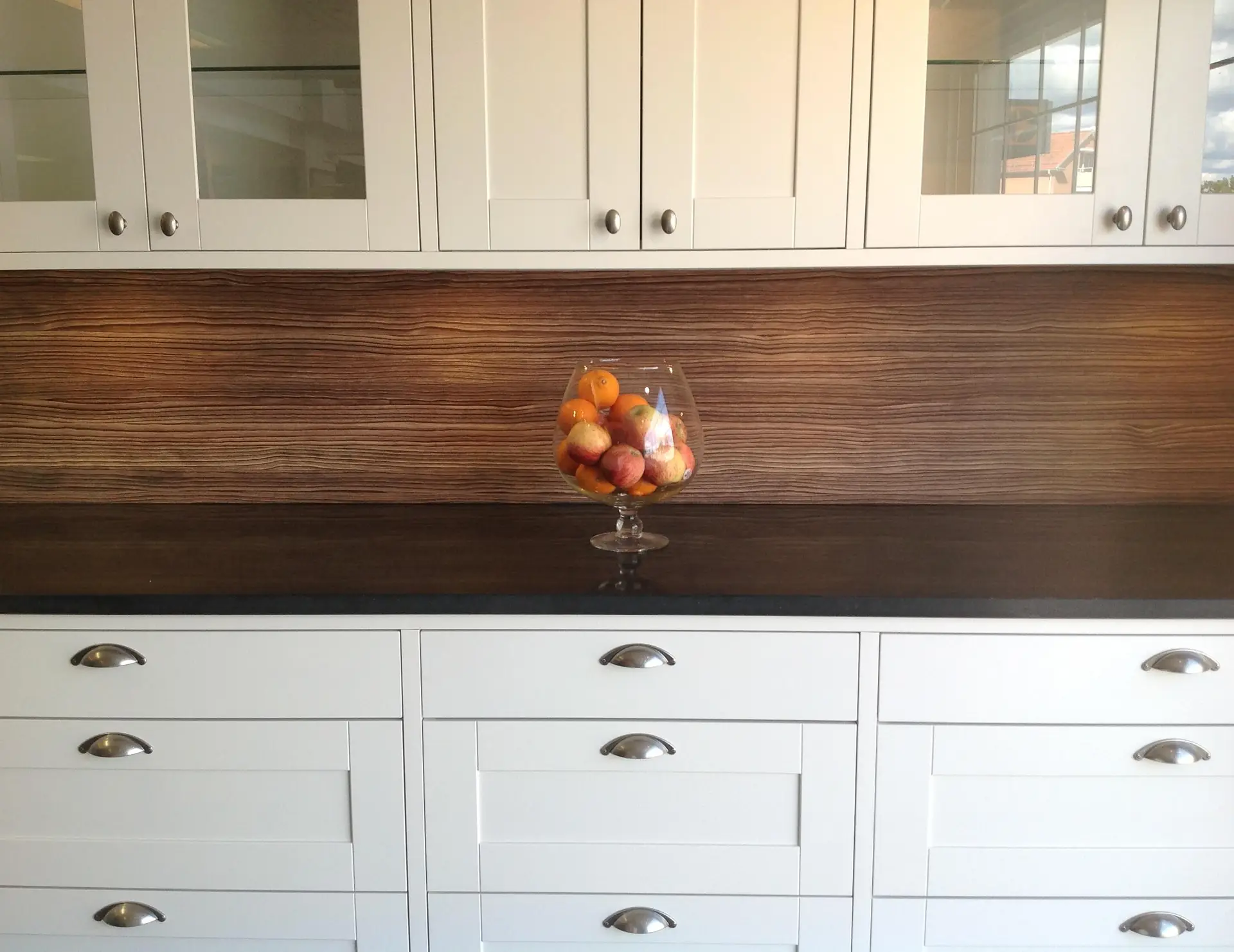 Luxury closet with custom white cabinetry, featuring elegant metallic handles and a dark countertop. A glass bowl with colorful fruit enhances the sophisticated ambiance against a wood-patterned backsplash.
