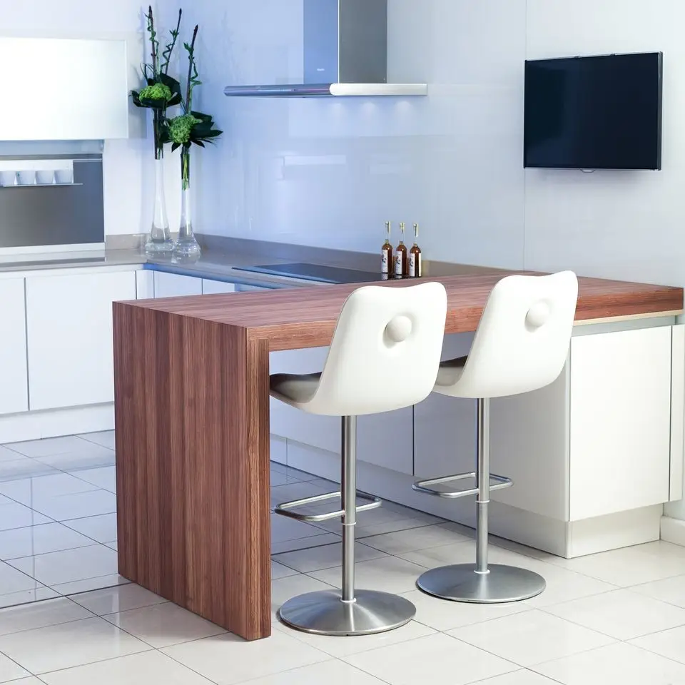 Modern kitchen design featuring custom cabinetry, with a sleek wood counter and two white bar stools. Elegant space with glossy white surfaces and decorative green plants.