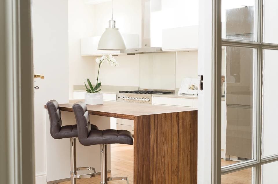 Modern kitchen design with custom cabinetry and luxury finishes, featuring a wooden island with two cushioned bar stools, pendant light, sleek appliances, and elegant decor.