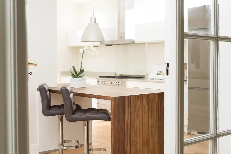 Modern kitchen design with custom cabinetry and luxury finishes, featuring a wooden island with two cushioned bar stools, pendant light, sleek appliances, and elegant decor.