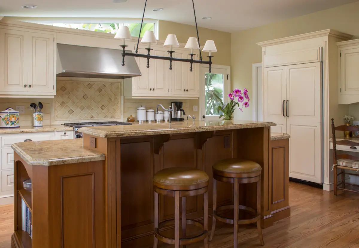 A luxury kitchen featuring custom cabinetry with elegant design. The space includes a central island with bar stools, polished granite countertops, and a decorative light fixture. The cabinetry is painted in a classic cream color, complemented by warm wood accents and a tiled backsplash. The design exudes sophistication and functionality, ideal for elegant kitchen spaces.