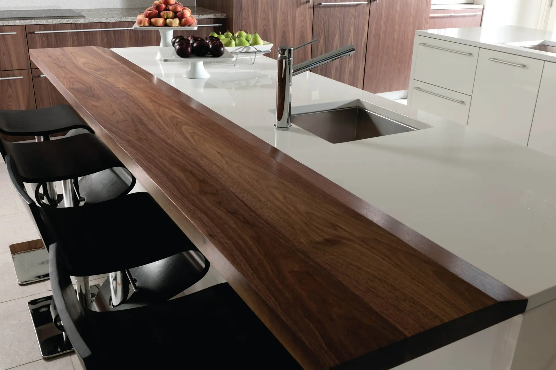 Luxurious kitchen design featuring a high-end island with a sleek faucet and integrated sink. The island combines white and wooden surfaces, paired with modern black bar stools. Surrounding cabinets have a transitional style, blending contemporary and traditional elements. A bowl of fresh fruits sits on the countertop, enhancing the sophisticated atmosphere.