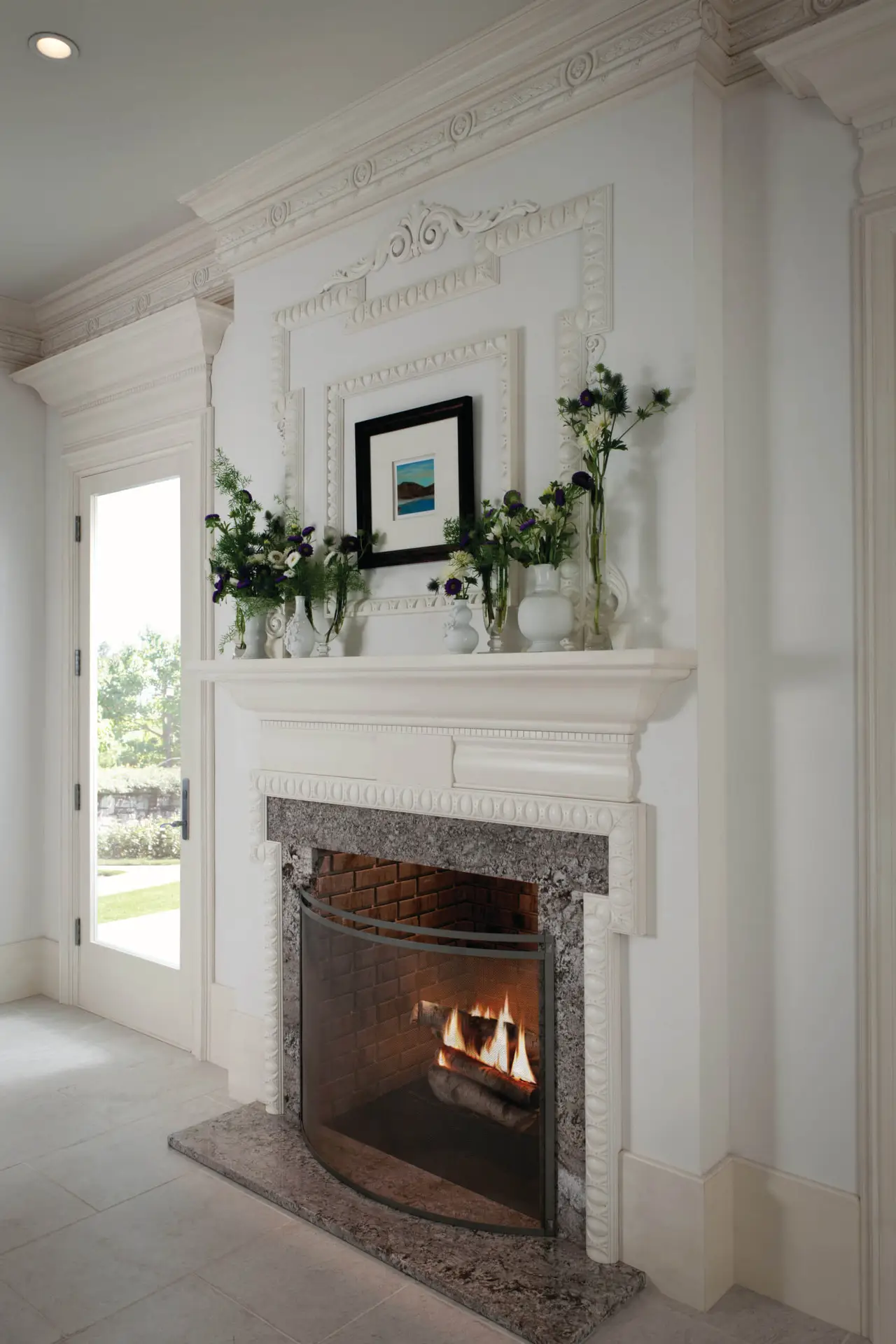 Elegant living room detail showcasing a luxurious white fireplace with intricate molding and a decorative mirror above. The mantel is adorned with green foliage and white flowers in vases, creating a classic and sophisticated ambiance.