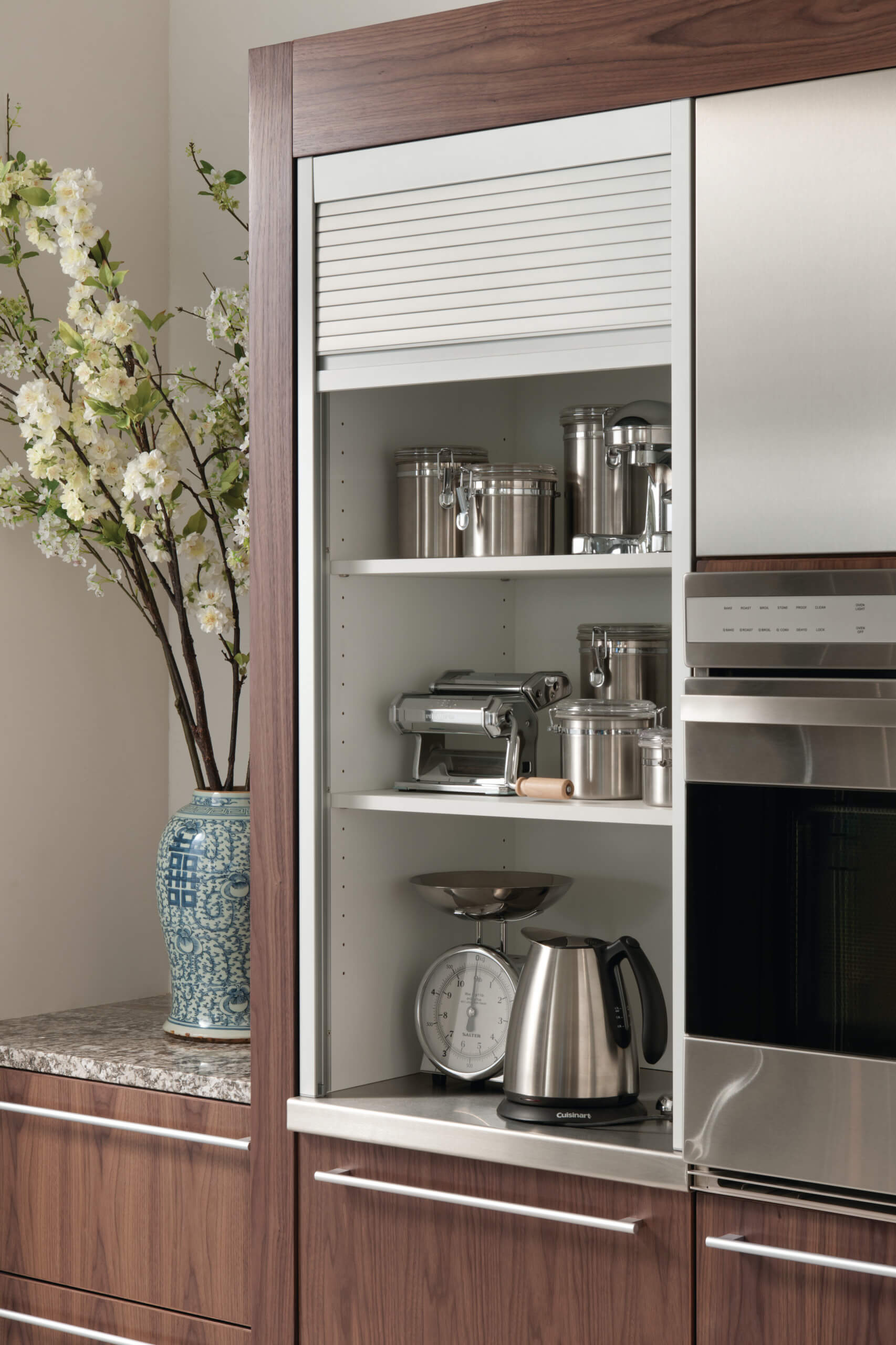 Luxury kitchen cabinet with transitional design, featuring sleek stainless steel appliances, a kettle and storage containers inside. A blue and white decorative vas