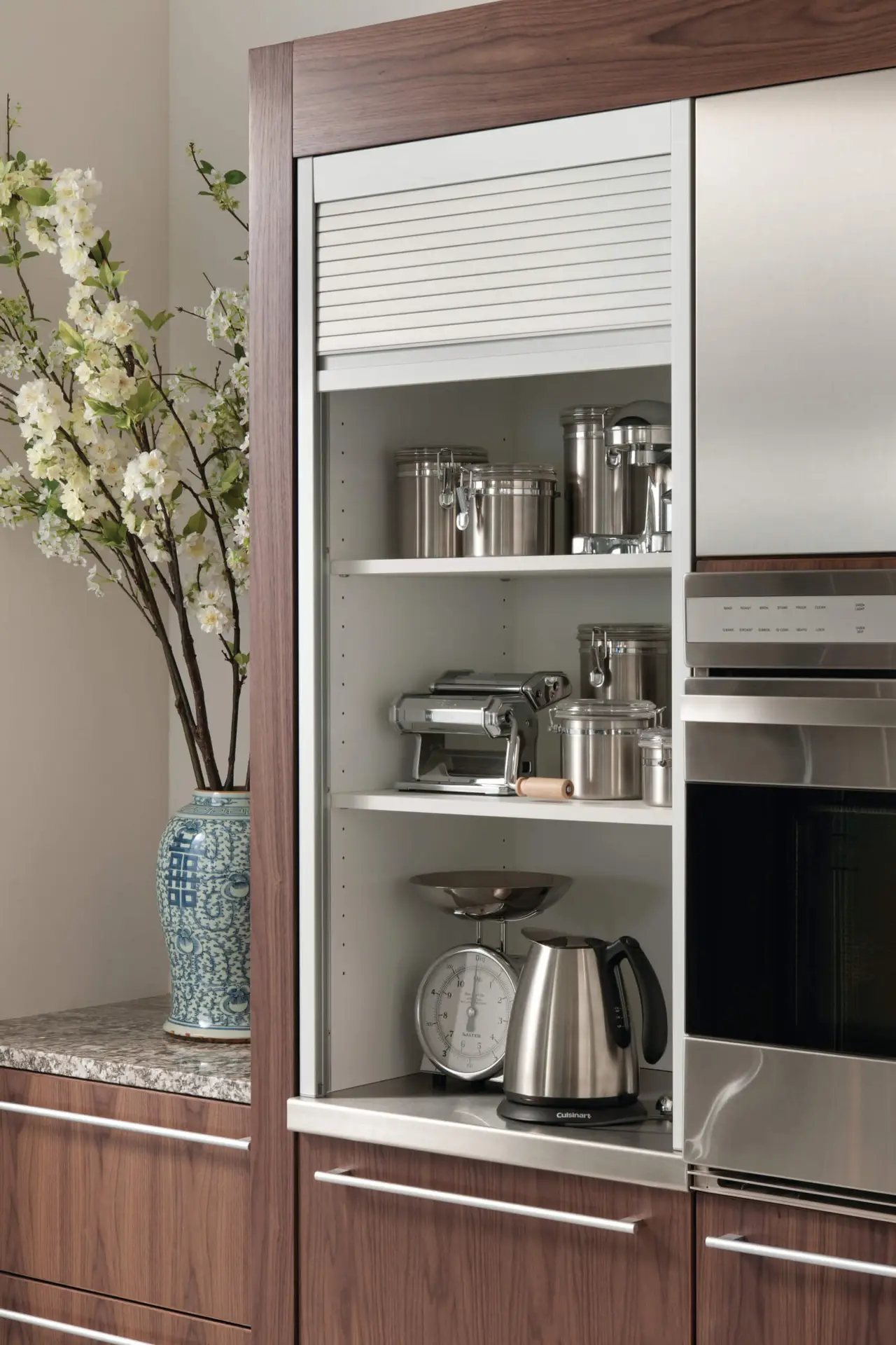 Luxury kitchen cabinet with transitional design, featuring sleek stainless steel appliances, a kettle and storage containers inside. A blue and white decorative vas