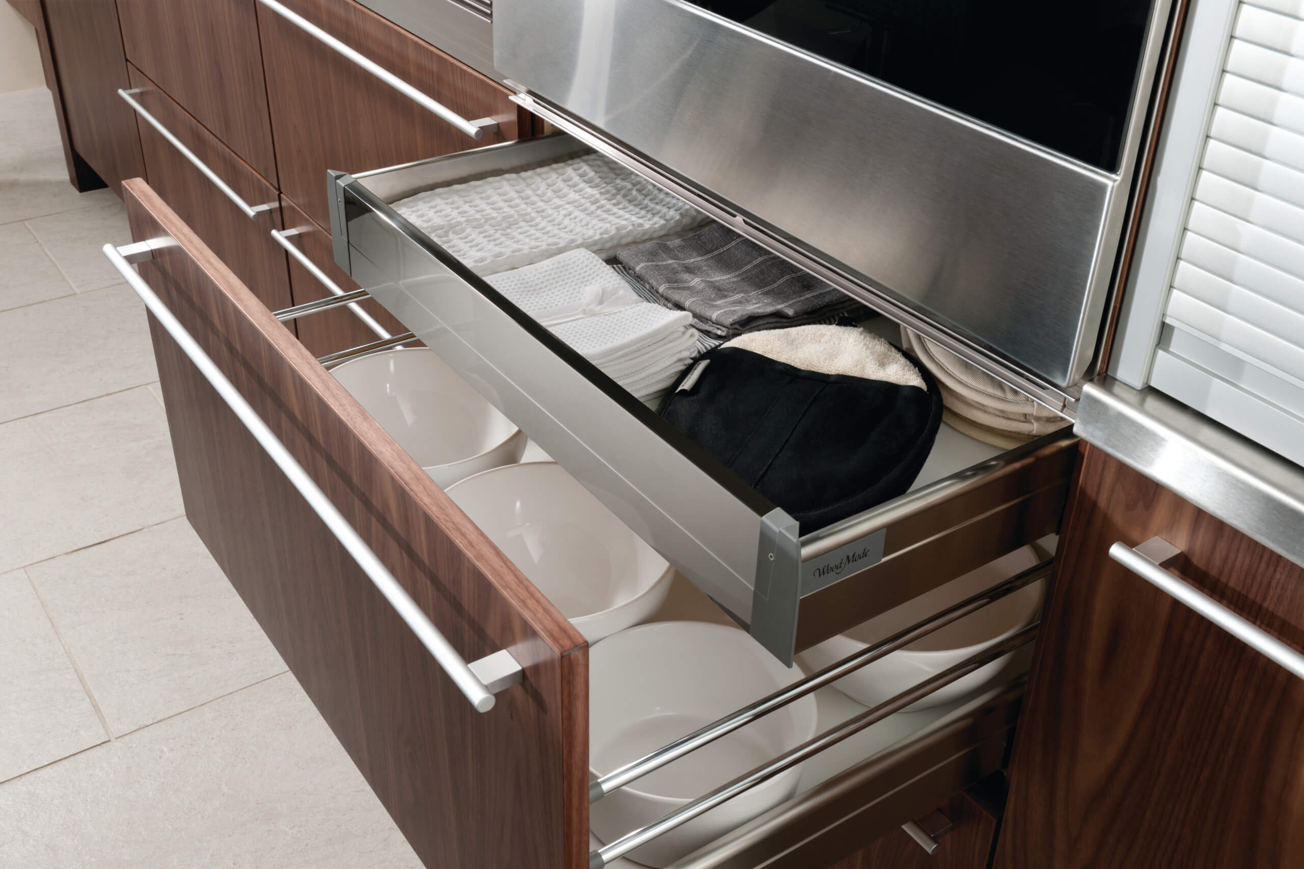 Modern luxury kitchen featuring transitional walnut cabinetry with sleek, horizontal metal handles. Open drawers reveal neatly organized kitchenware and folded linens, exemplifying sophisticated kitchen storage solutions.
