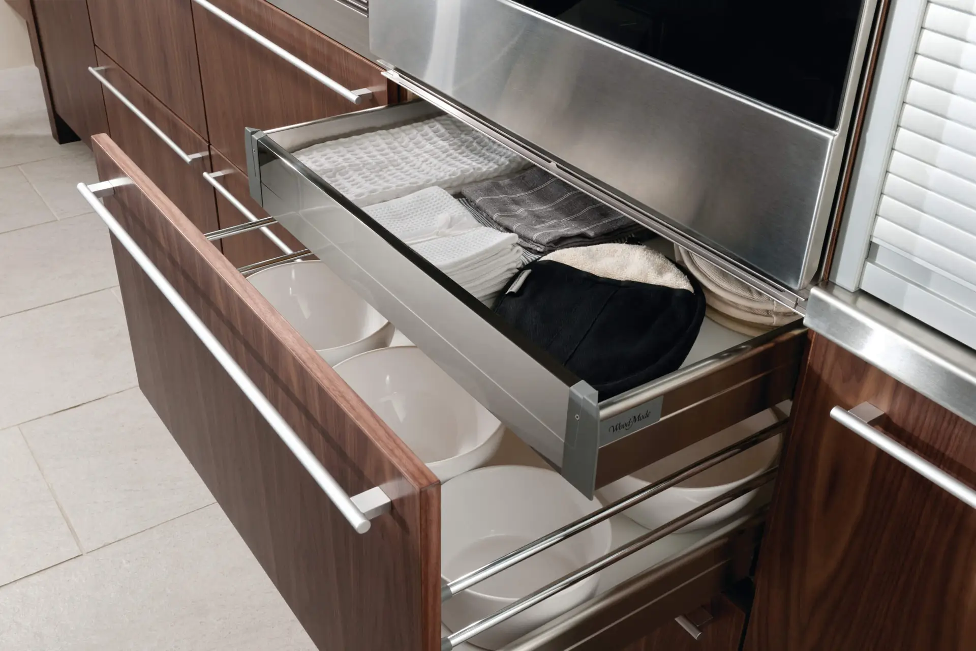 Modern luxury kitchen featuring transitional walnut cabinetry with sleek, horizontal metal handles. Open drawers reveal neatly organized kitchenware and folded linens, exemplifying sophisticated kitchen storage solutions.