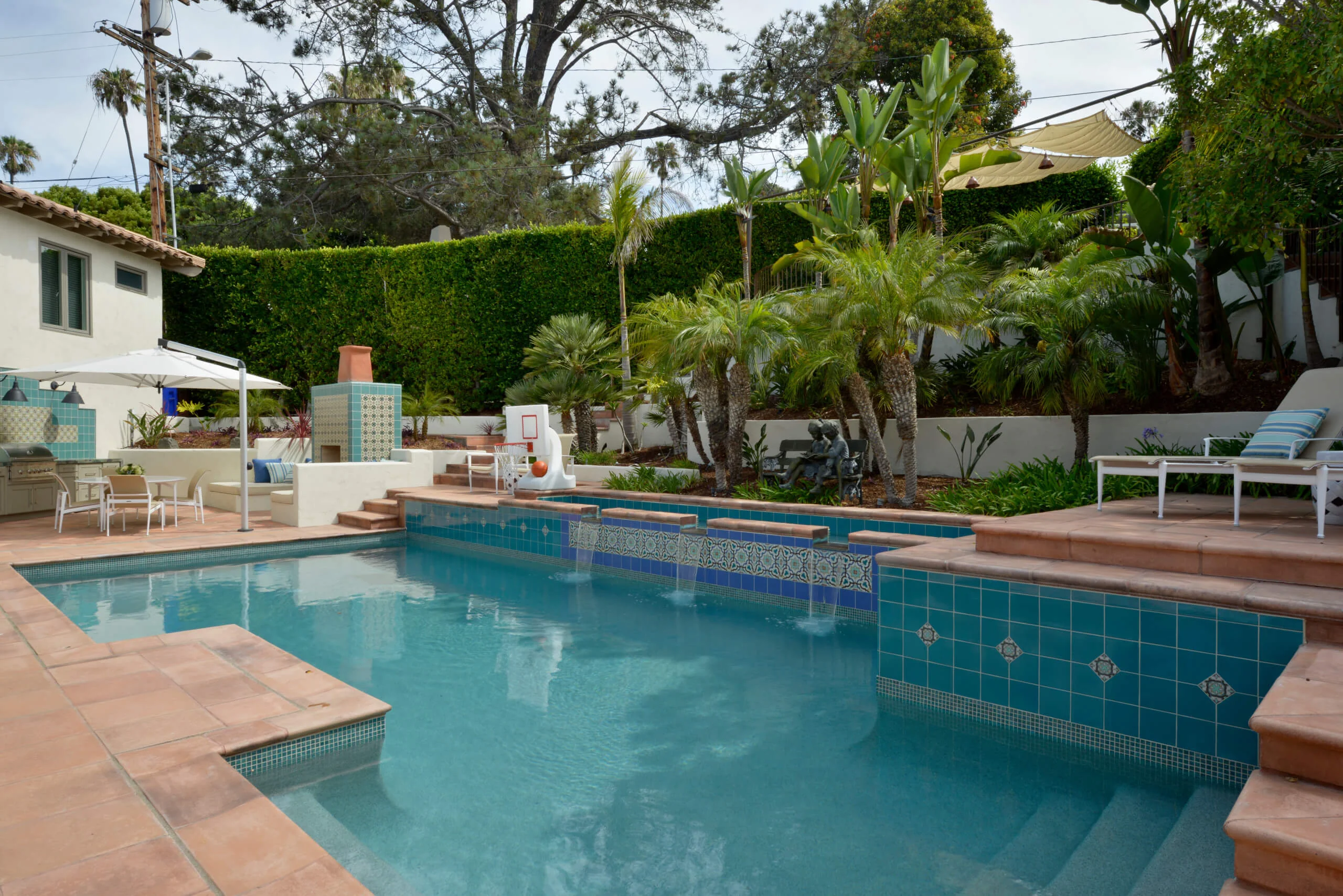 Spacious backyard featuring a luxurious swimming pool with turquoise tiles, surrounded by lush greenery and palm trees