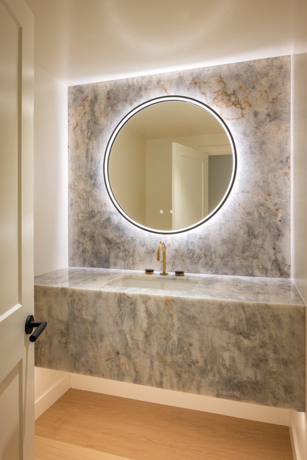 Floating natural stone powder room vanity with full-height stone slab wall and backlit round mirror in La Jolla